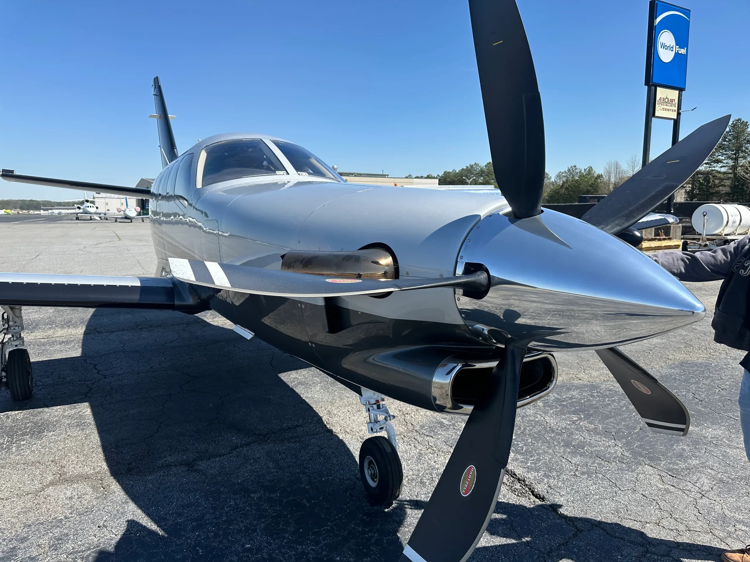 A front-right view of a silver and black TBM 940 aircraft with its propeller, parked on an asphalt tarmac.