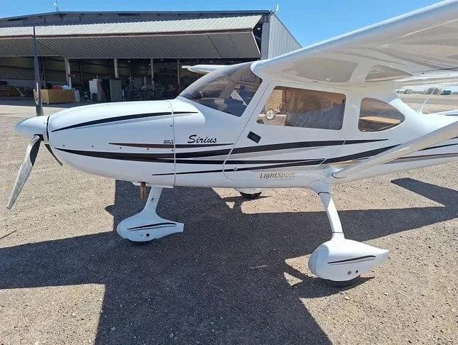 A close-up side view of the front of a white Sirius LightSport aircraft with black and grey stripes, showing the propeller and nose.
