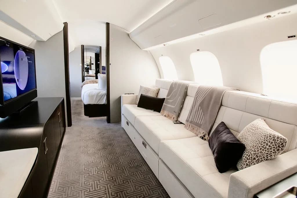 A luxurious private jet interior featuring a long white sofa with throw pillows, a television, and a glimpse of a bedroom area.