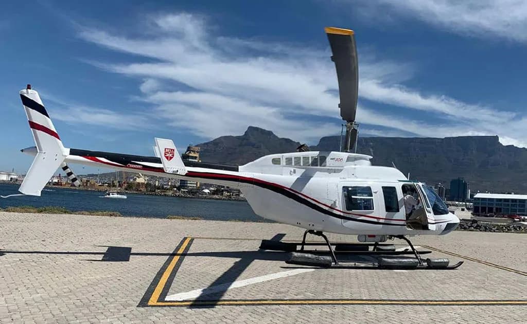 A white helicopter with black and red stripes on a helipad with a large mountain and city in the background.