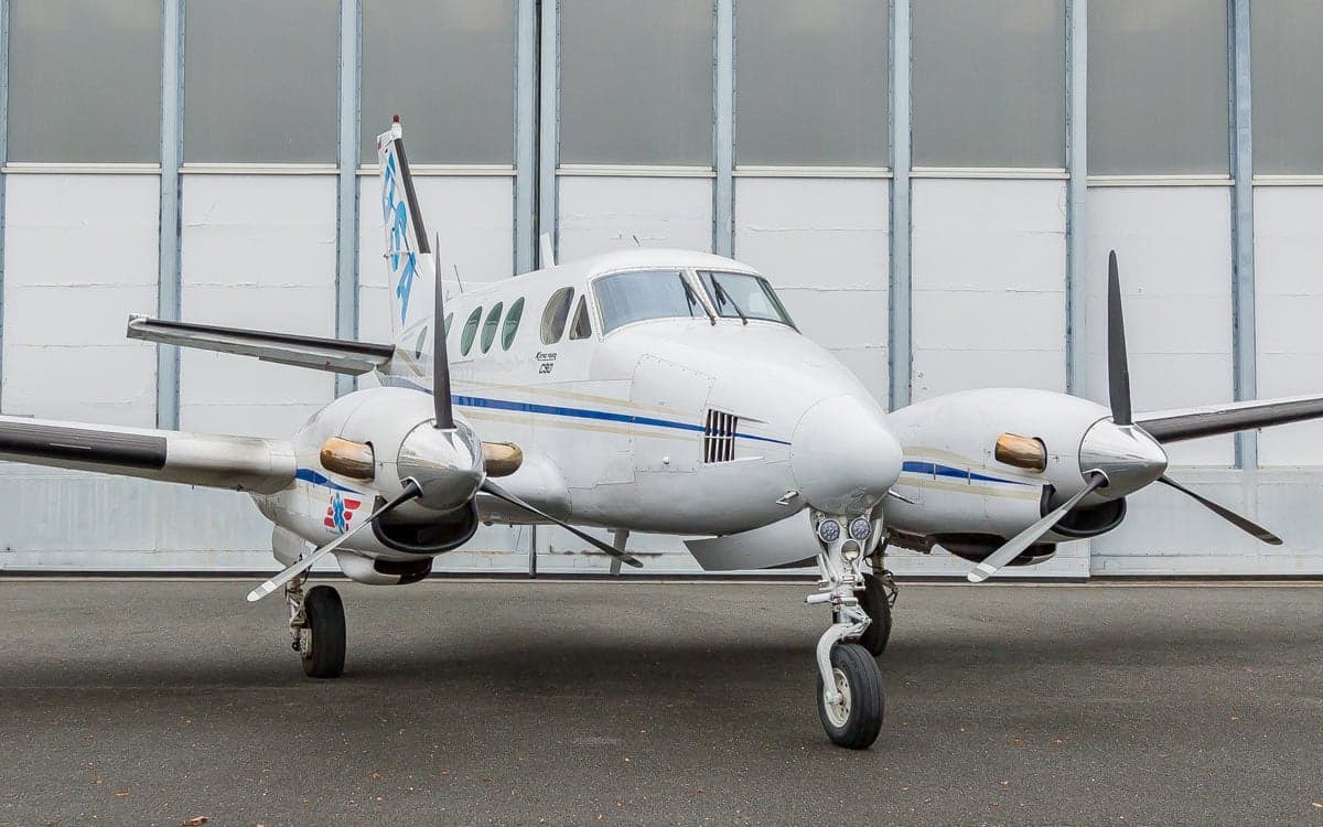 Front view of a Beechcraft King Air C90 aircraft with a white and blue paint scheme, parked in front of a hangar.