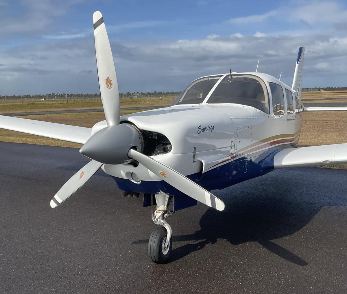 A close-up front view of a white and blue Piper Saratoga aircraft showing its propeller and nose on a wet tarmac.