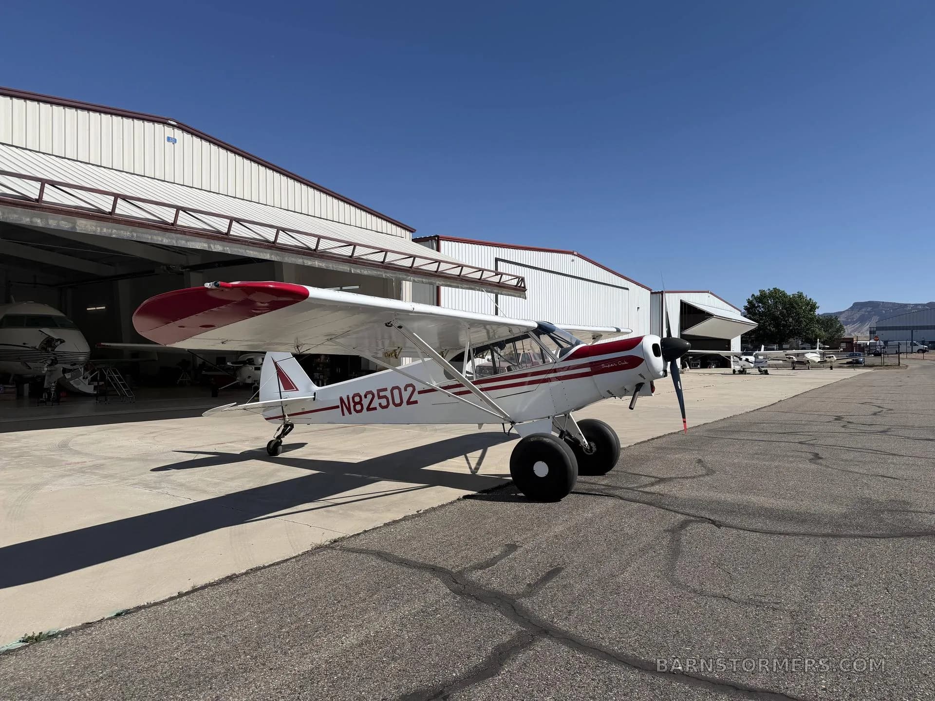A white and red single-engine bush plane with large tires is parked on an asphalt apron in front of hangars under a clear blue sky.