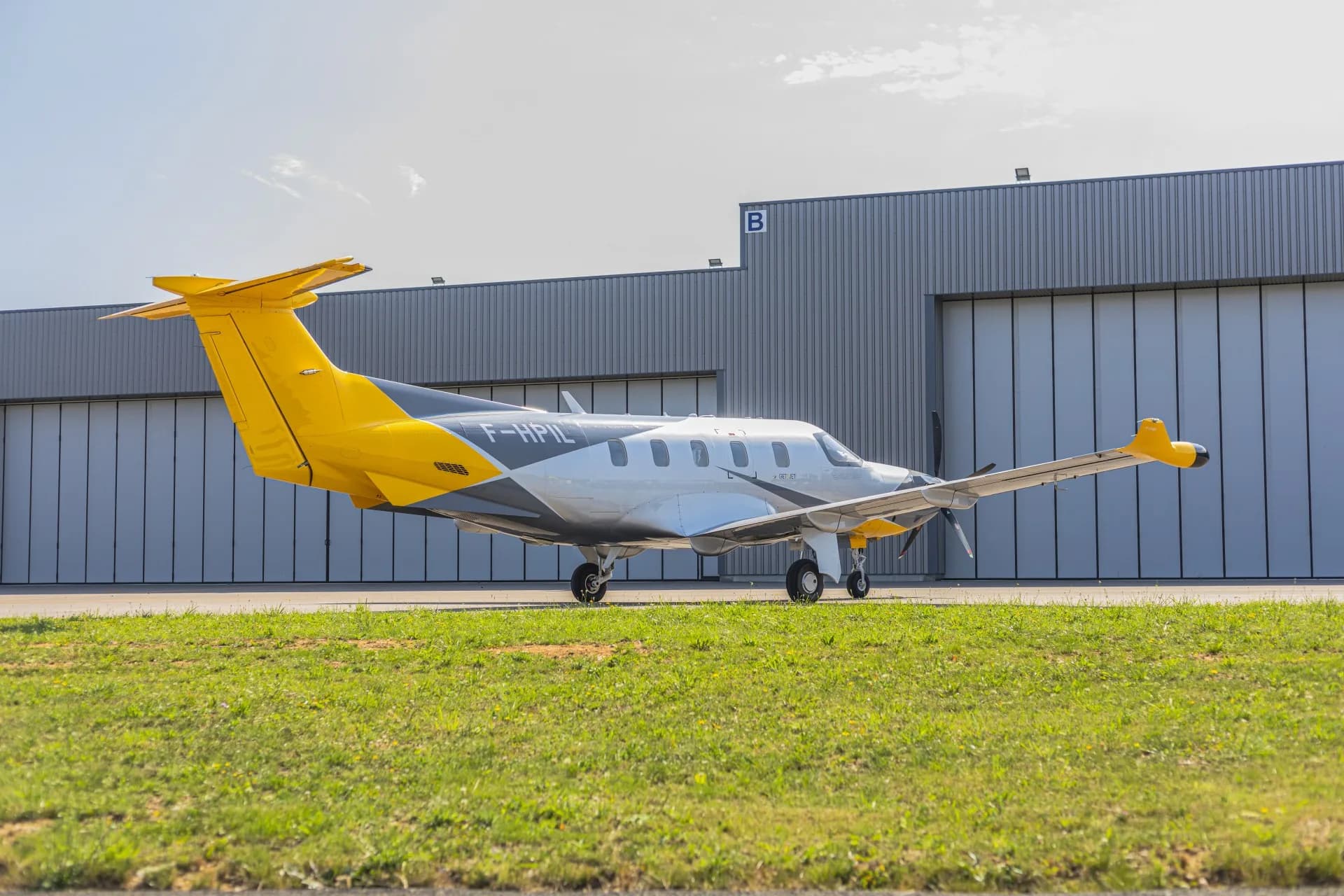 A silver and yellow Pilatus PC-12 aircraft (F-HPIL) parked on the tarmac next to a large hangar.