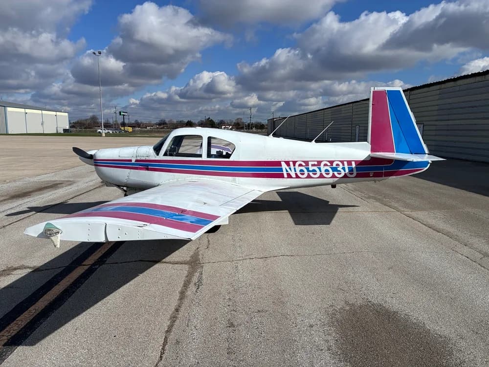 A Piper PA-28-140 Cherokee with a white, blue, and pink paint scheme on the tarmac.