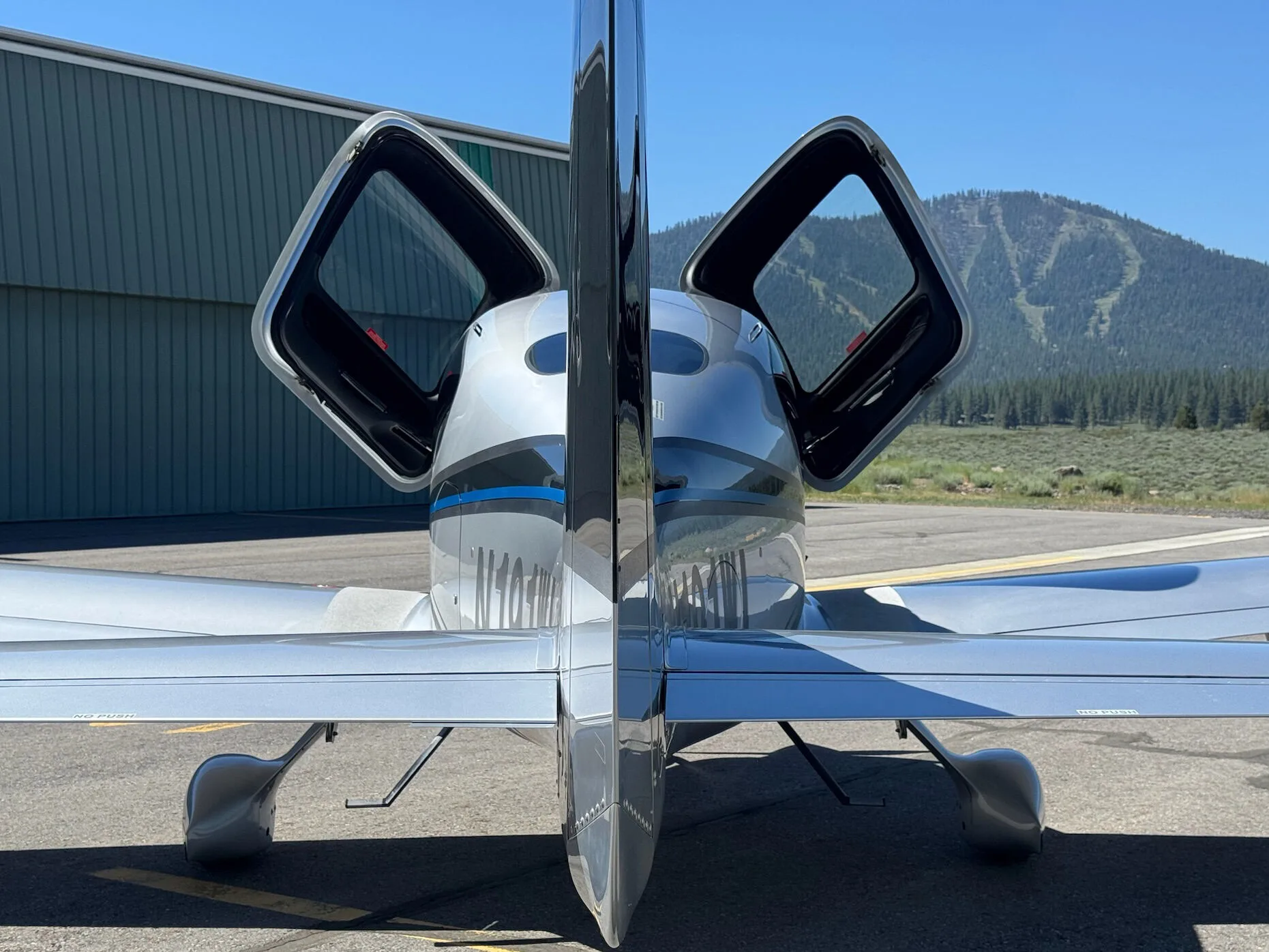 A rear view of a silver and black Cirrus SR22T aircraft with N191WL, showing the tail and both doors open, with mountains in the background.