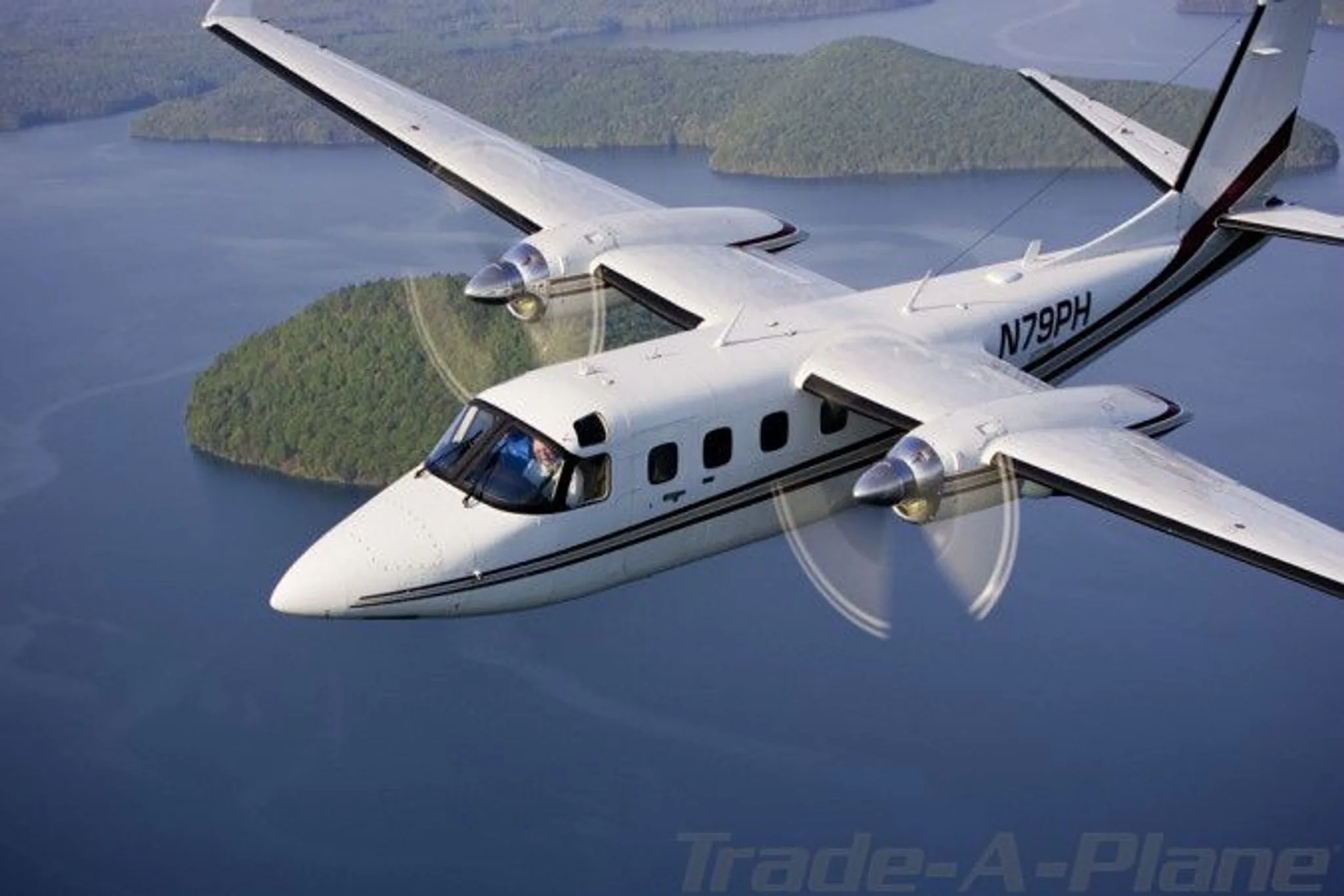 A white twin-engine turboprop aircraft, N79PH, with black and red stripes, flying over a body of water with green islands.