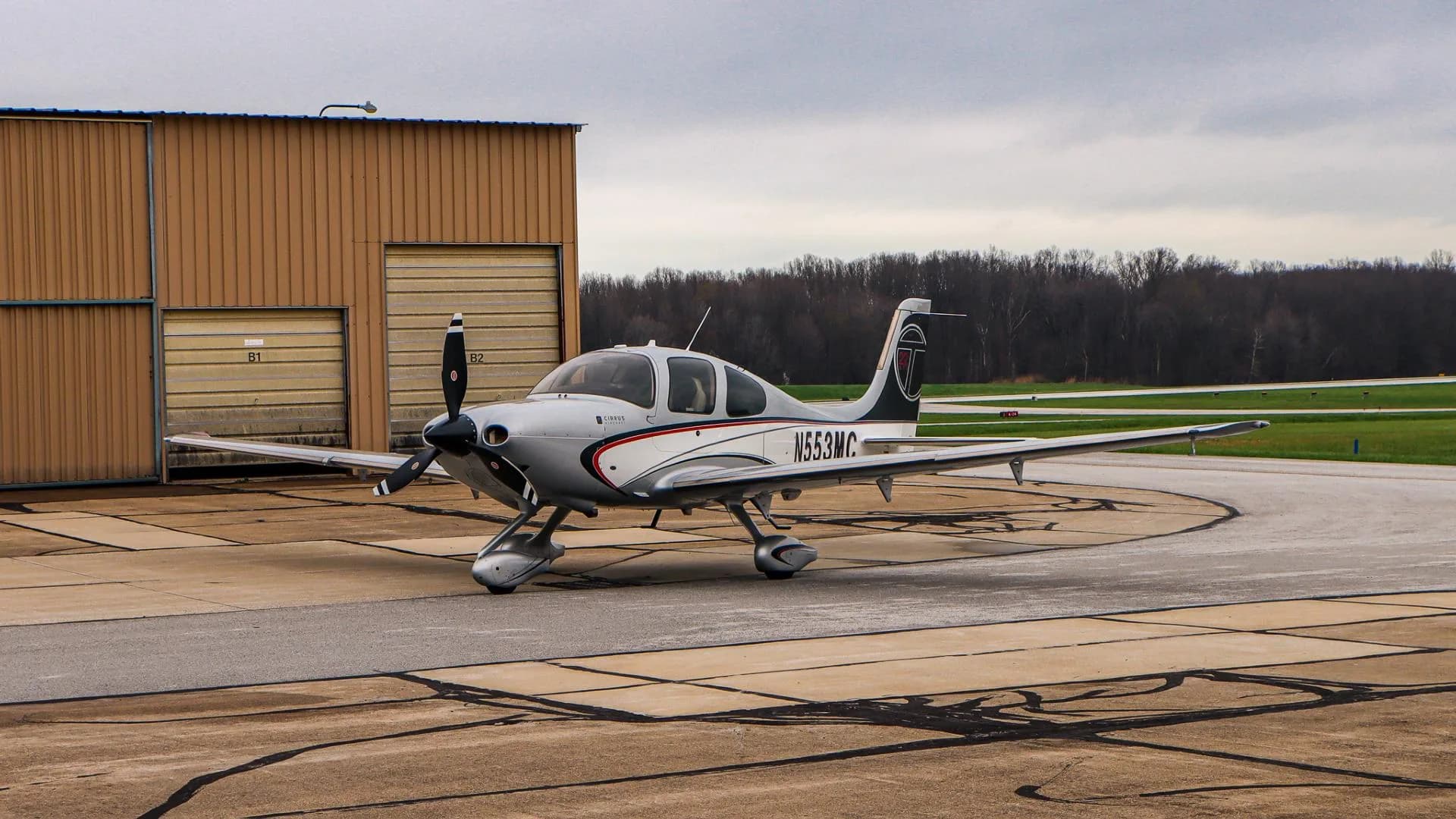 A silver and white Cirrus SR22 aircraft with N553MC registration is parked on an airport tarmac next to a hangar.