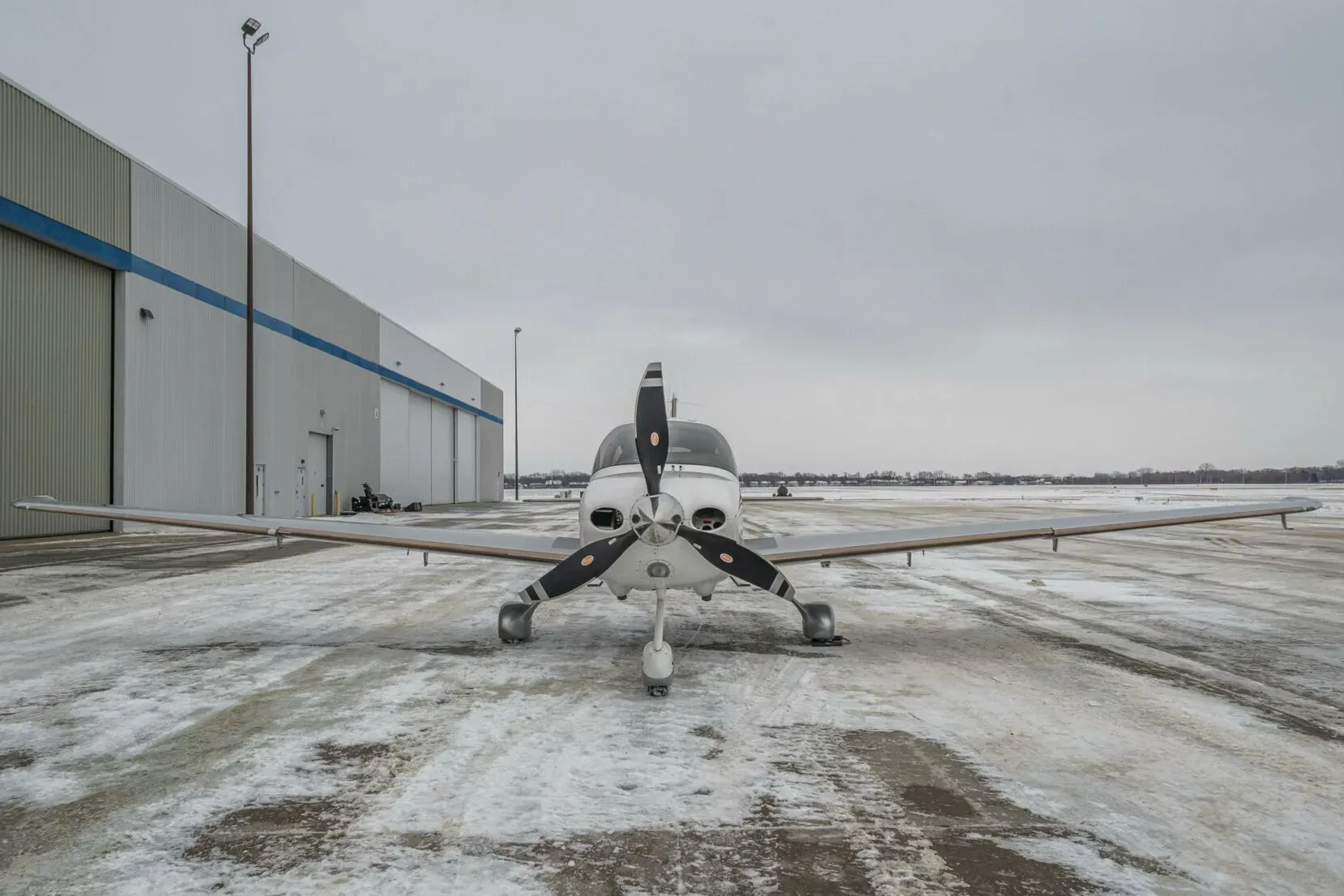A white and grey Cirrus SR22T aircraft (C-GWHA) viewed from the front, parked on a snowy tarmac in front of a hangar.