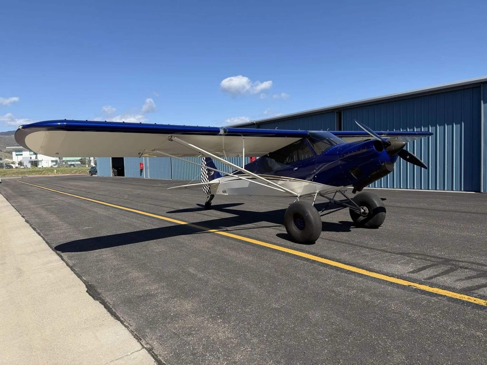 A blue and white Carbon Cub SS aircraft with large tundra tires, seen from the front-left side on an airport tarmac.