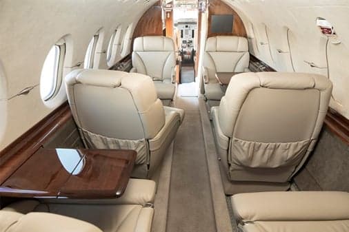 A view looking forward down the aisle of an aircraft cabin with light leather seats and a wooden table.