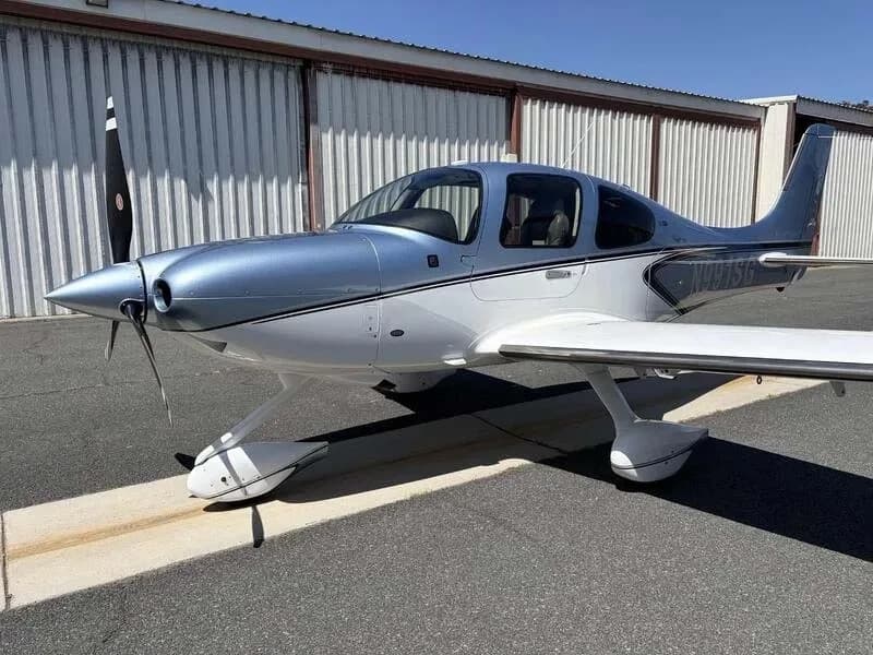 A blue and white Cirrus SR22 aircraft is parked on an asphalt ramp in front of a hangar.