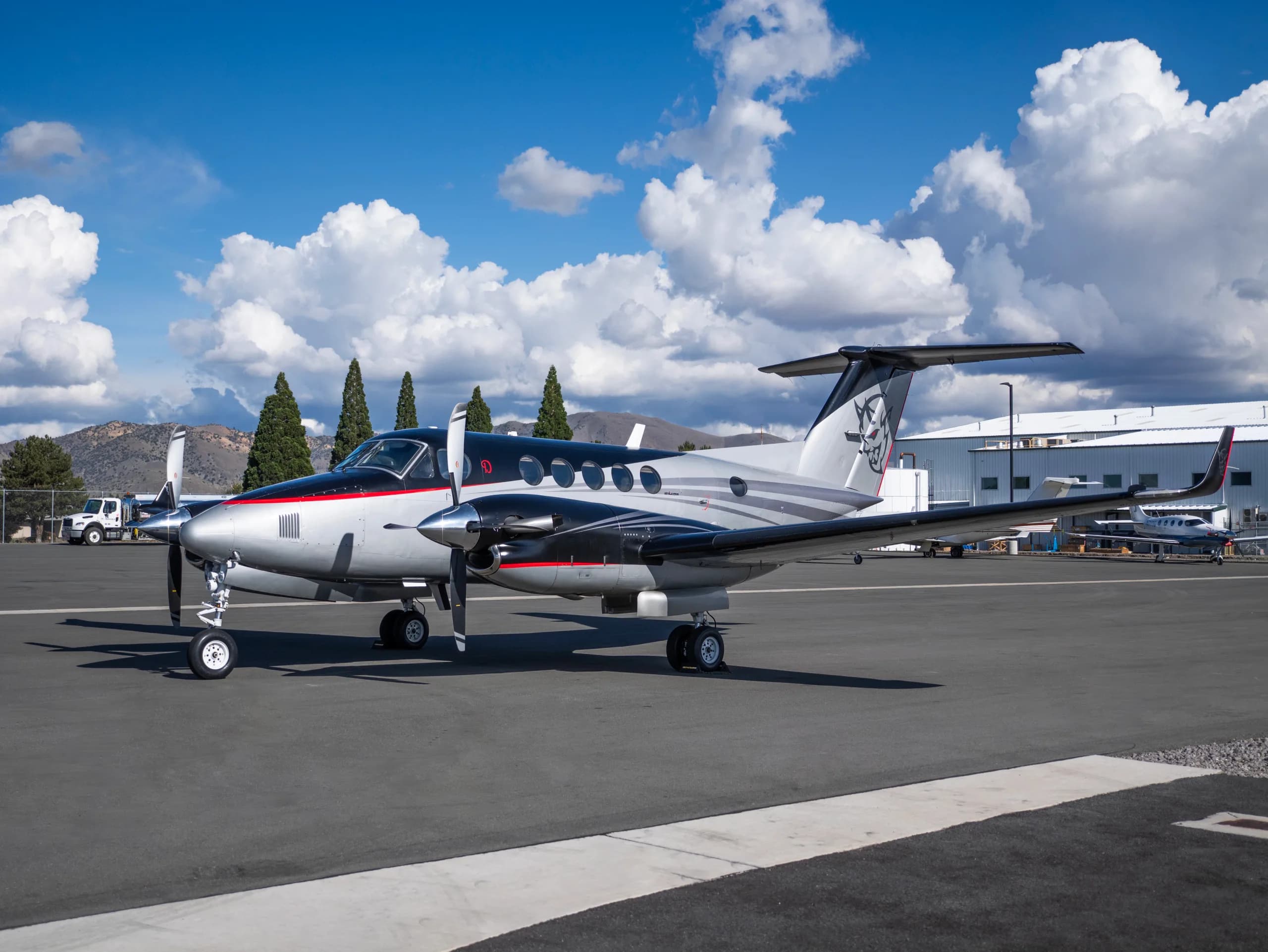 A silver and black twin-turboprop aircraft with red accents and a custom tail logo parked on an airport tarmac under a cloudy sky.