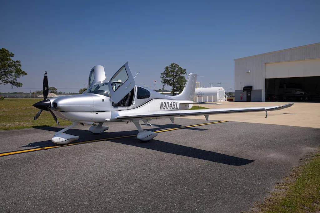 A front three-quarter view of the silver Cirrus SR22 aircraft N904BL with its doors open, parked in front of a hangar.