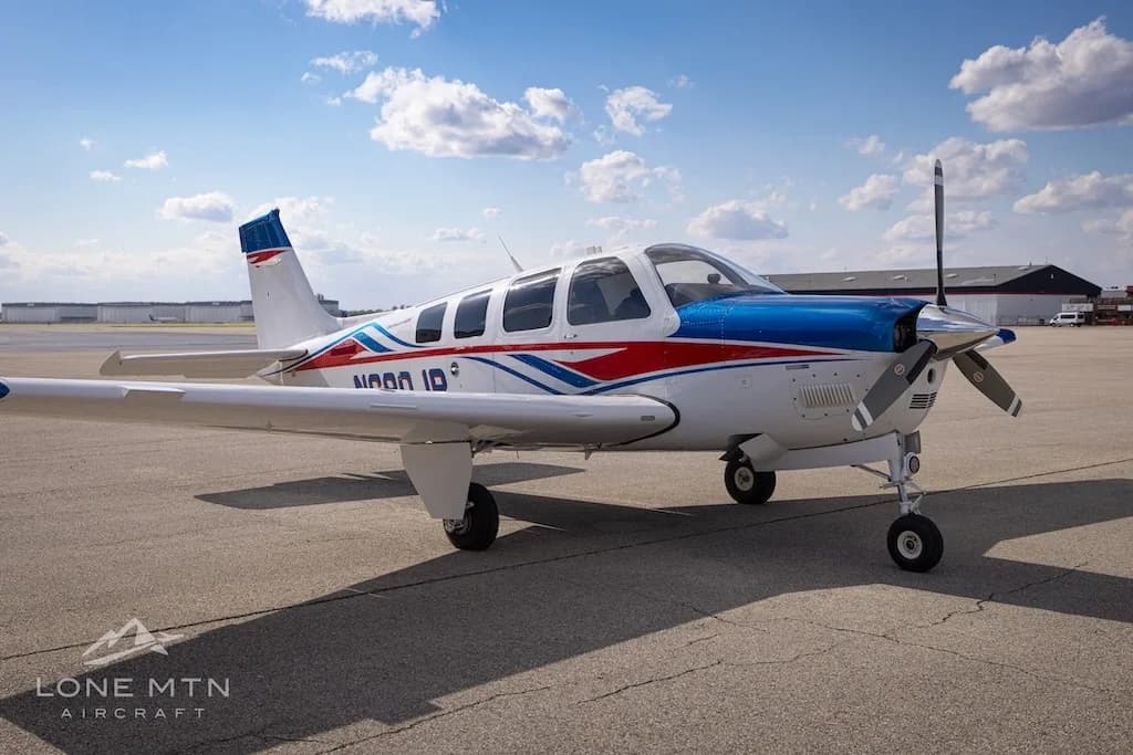 A white, blue, and red Beechcraft Bonanza N680JP parked on an airfield under a partly cloudy sky.