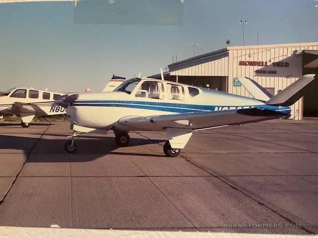 A white Beechcraft Bonanza with blue stripes is parked on a tarmac at Mondell Field, with another aircraft and a hangar in the background.