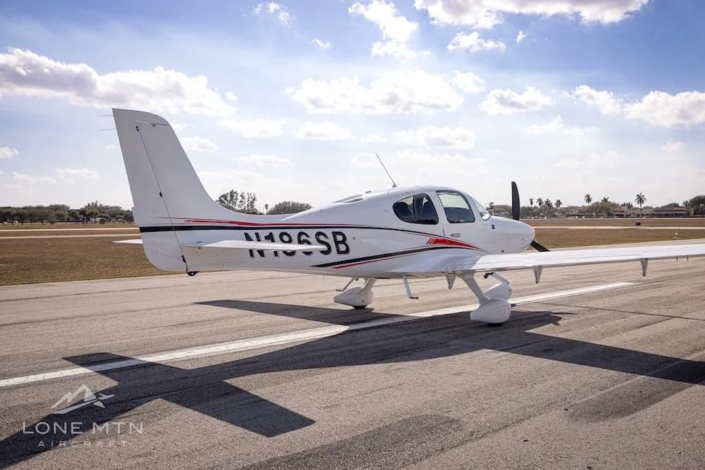 A rear-side view of the white N186SB single-engine aircraft with red and black stripes, parked on an airfield.