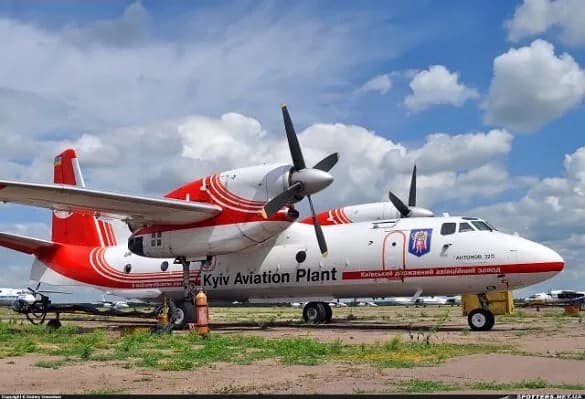 A white and red Antonov An-32B aircraft with 'Kyiv Aviation Plant' text on its fuselage, parked on a grassy tarmac under a partly cloudy sky.