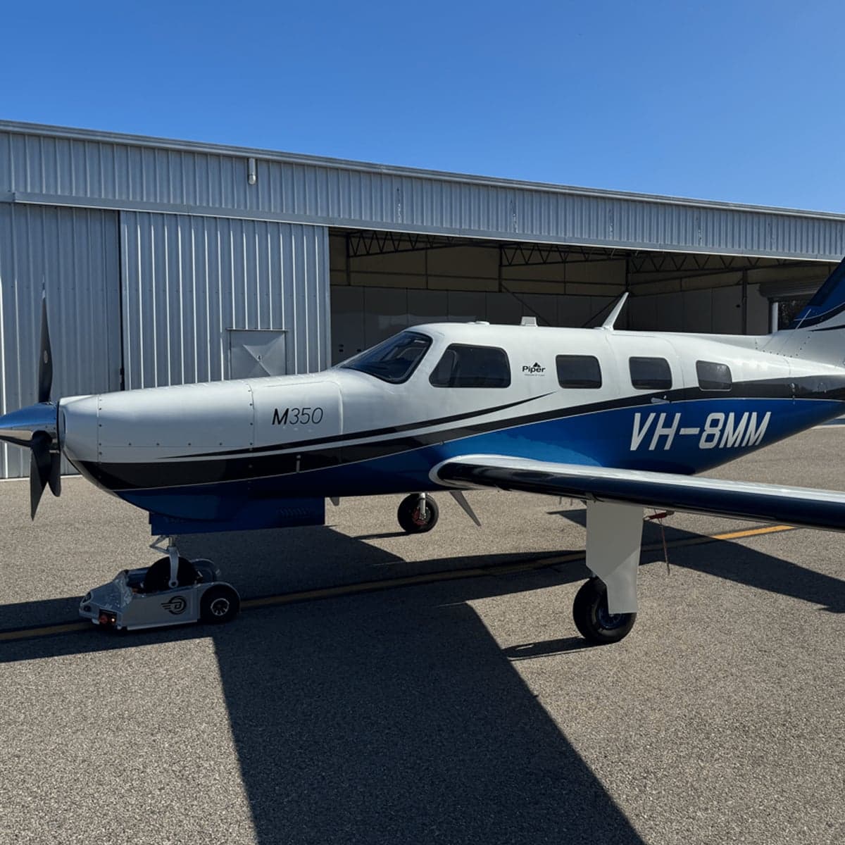 A white and blue Piper M350 aircraft parked on the tarmac in front of a hangar.