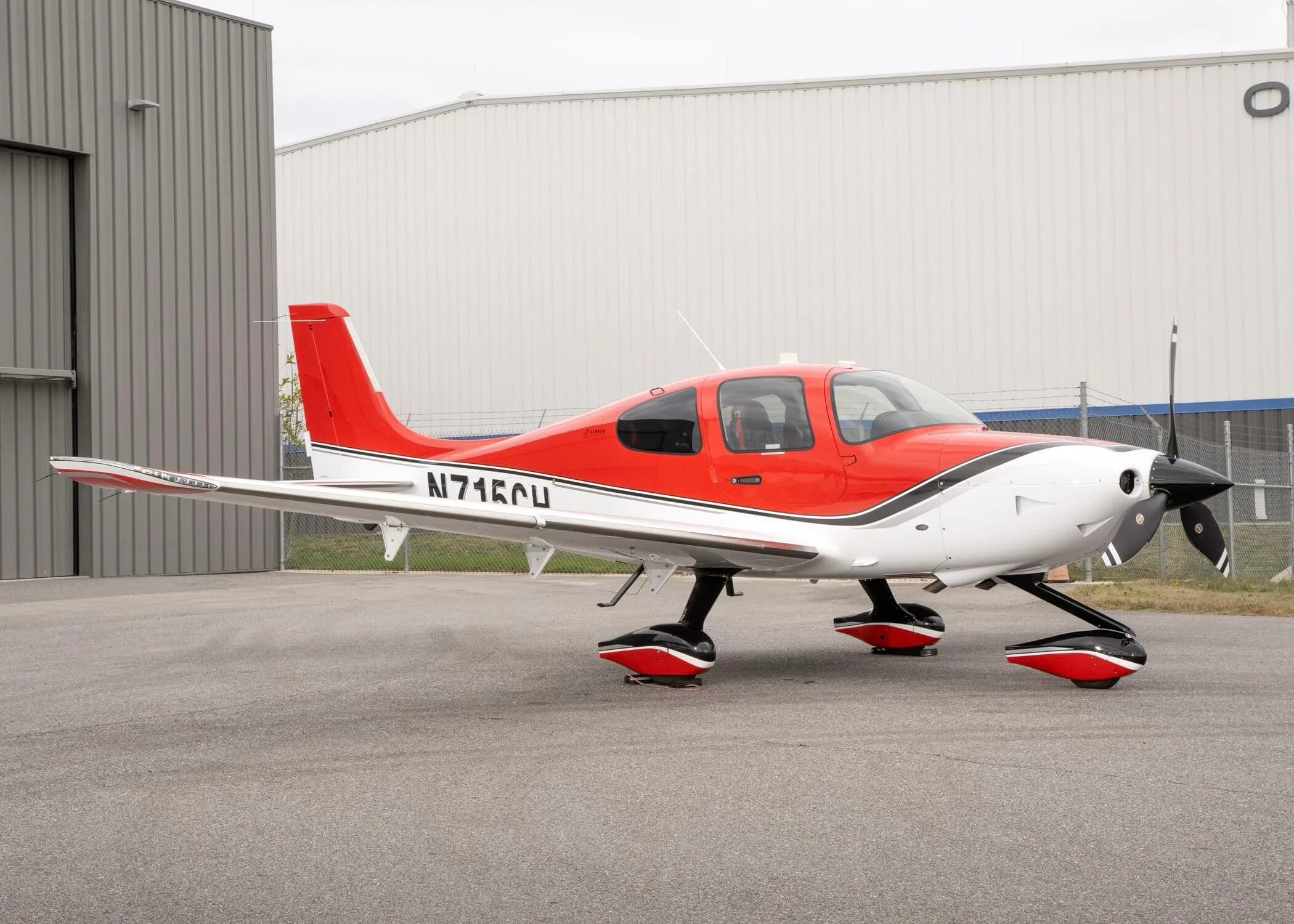 A red and white Cirrus SR22 aircraft with registration N715CH parked on an airfield next to a white hangar.