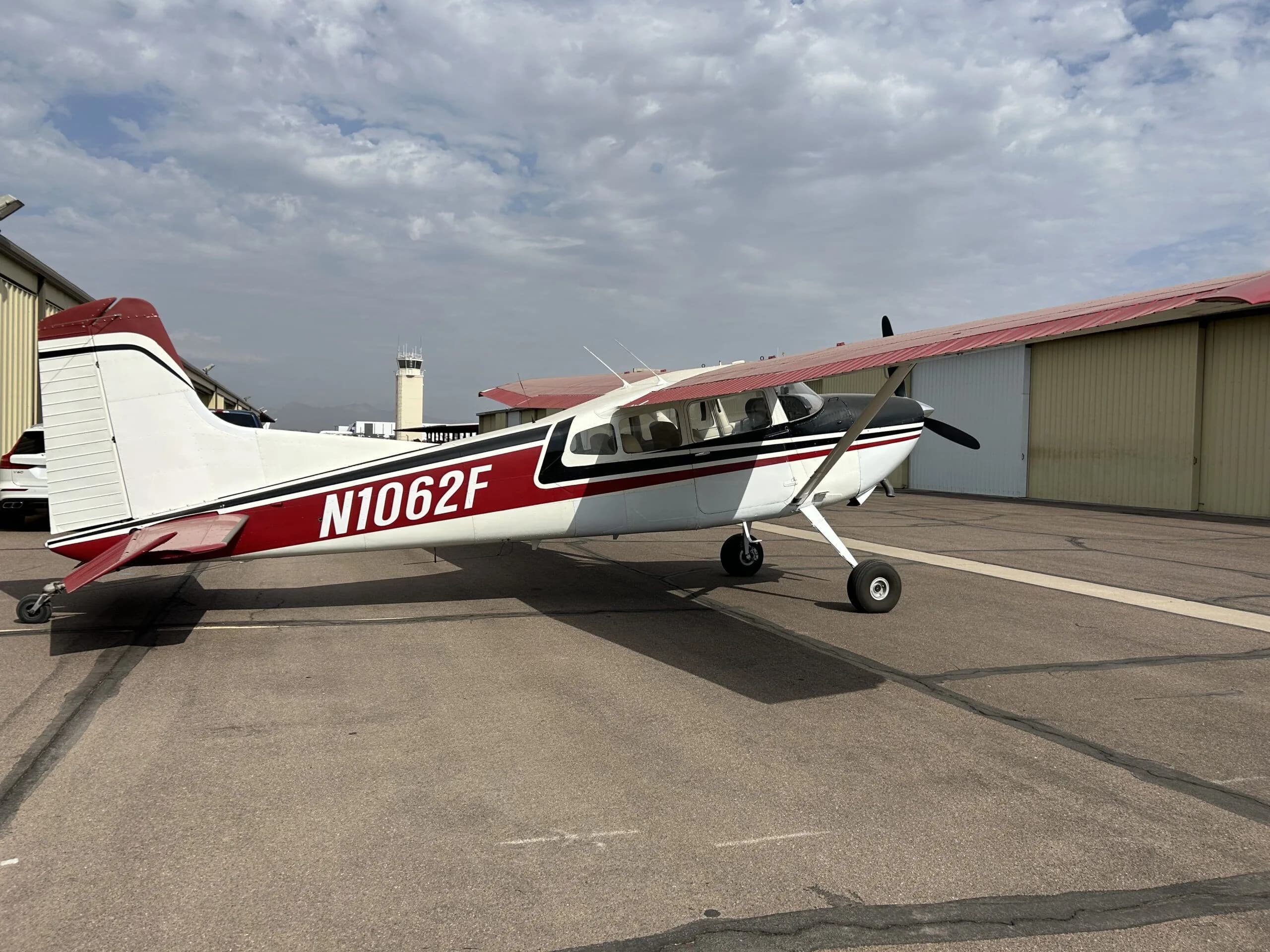 A white, black, and red Cessna 180 aircraft (N1062F) parked on an airport tarmac with an air traffic control tower in the distance.