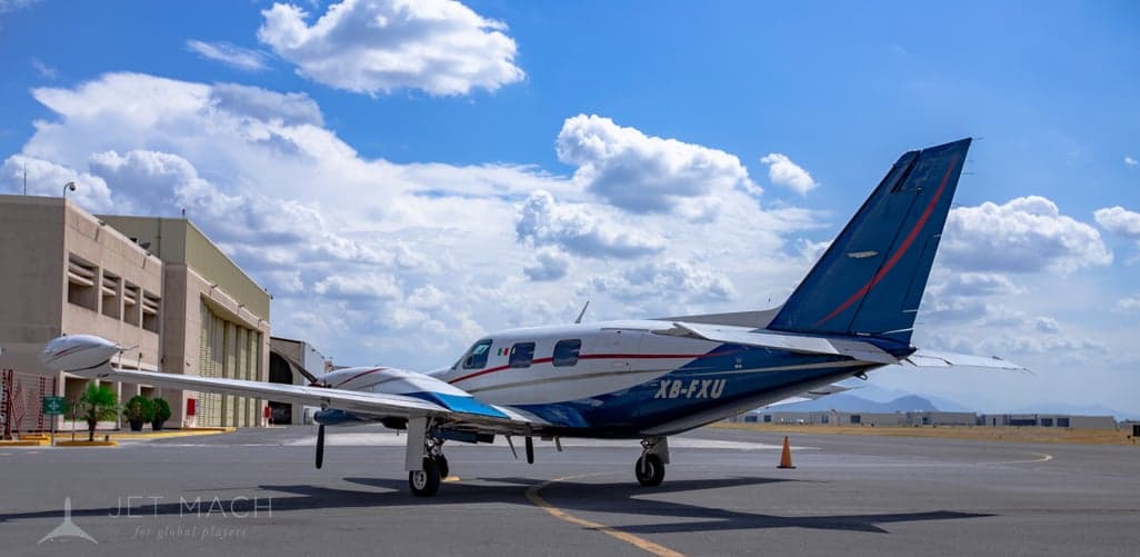A white and blue twin-engine propeller aircraft, registration XB-FXU, is parked on an airport tarmac with mountains in the distance.