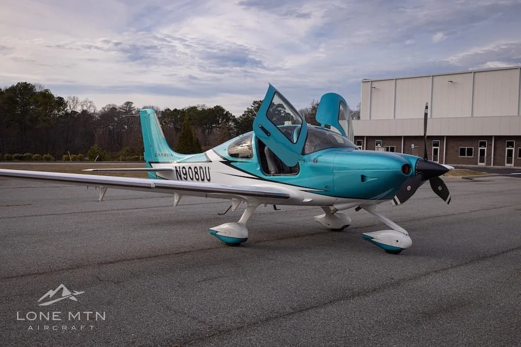 A front-side view of the teal and white Cirrus SR22 aircraft, N908DU, with its doors open, parked on an airfield.