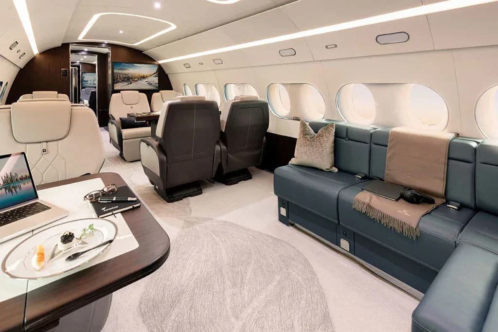 A private jet cabin featuring a blue leather sofa, individual seats, a laptop on a side table, and a TV screen.