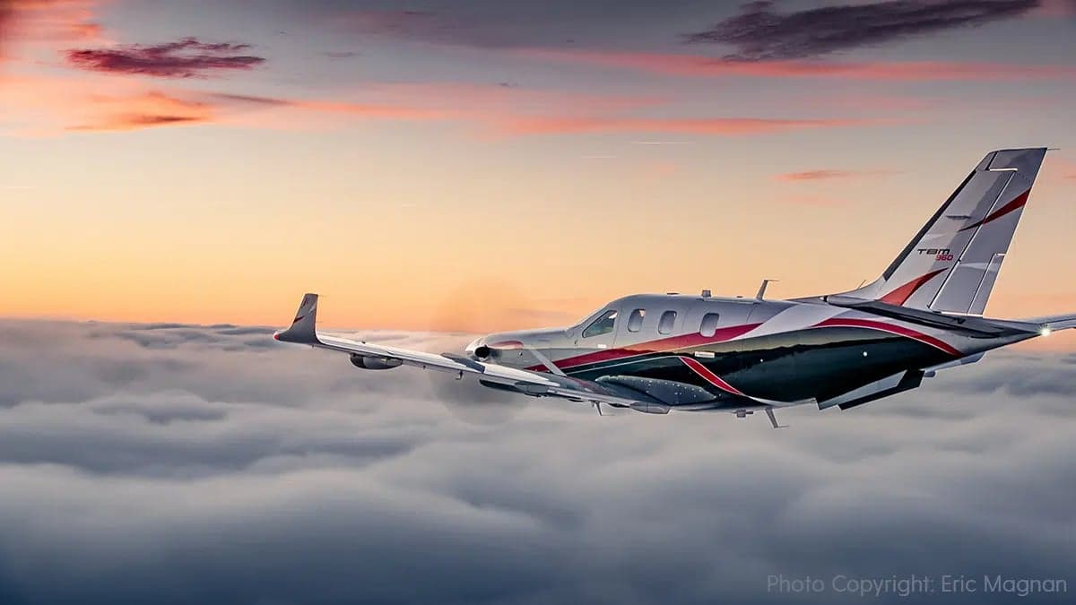 A TBM 960 aircraft with a silver, black, and red paint scheme flying above clouds at sunset.