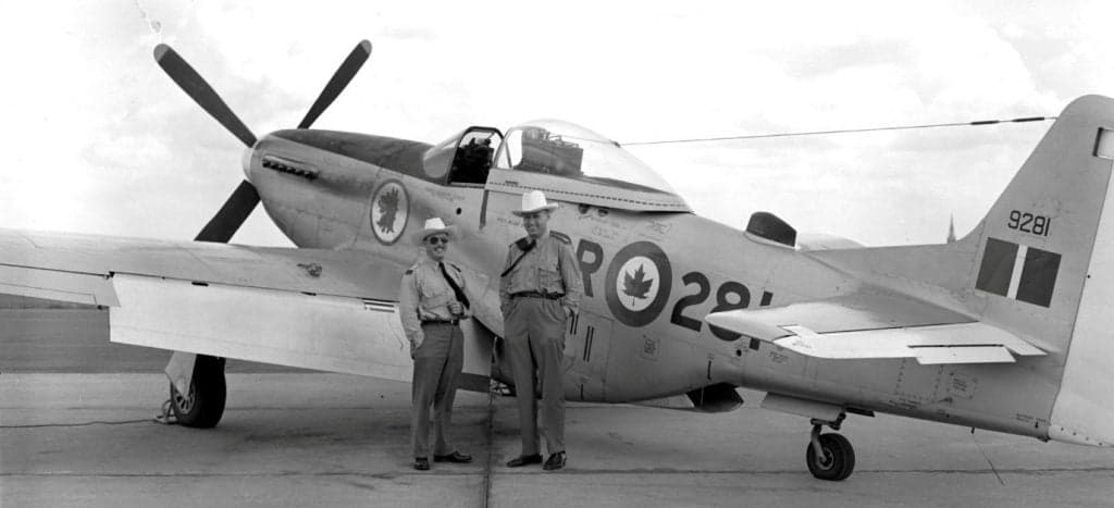 Two men pose in front of a vintage P-51 Mustang with Canadian markings.