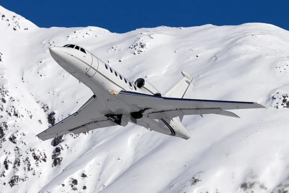 A white Falcon jet climbs over a snowy mountain range.