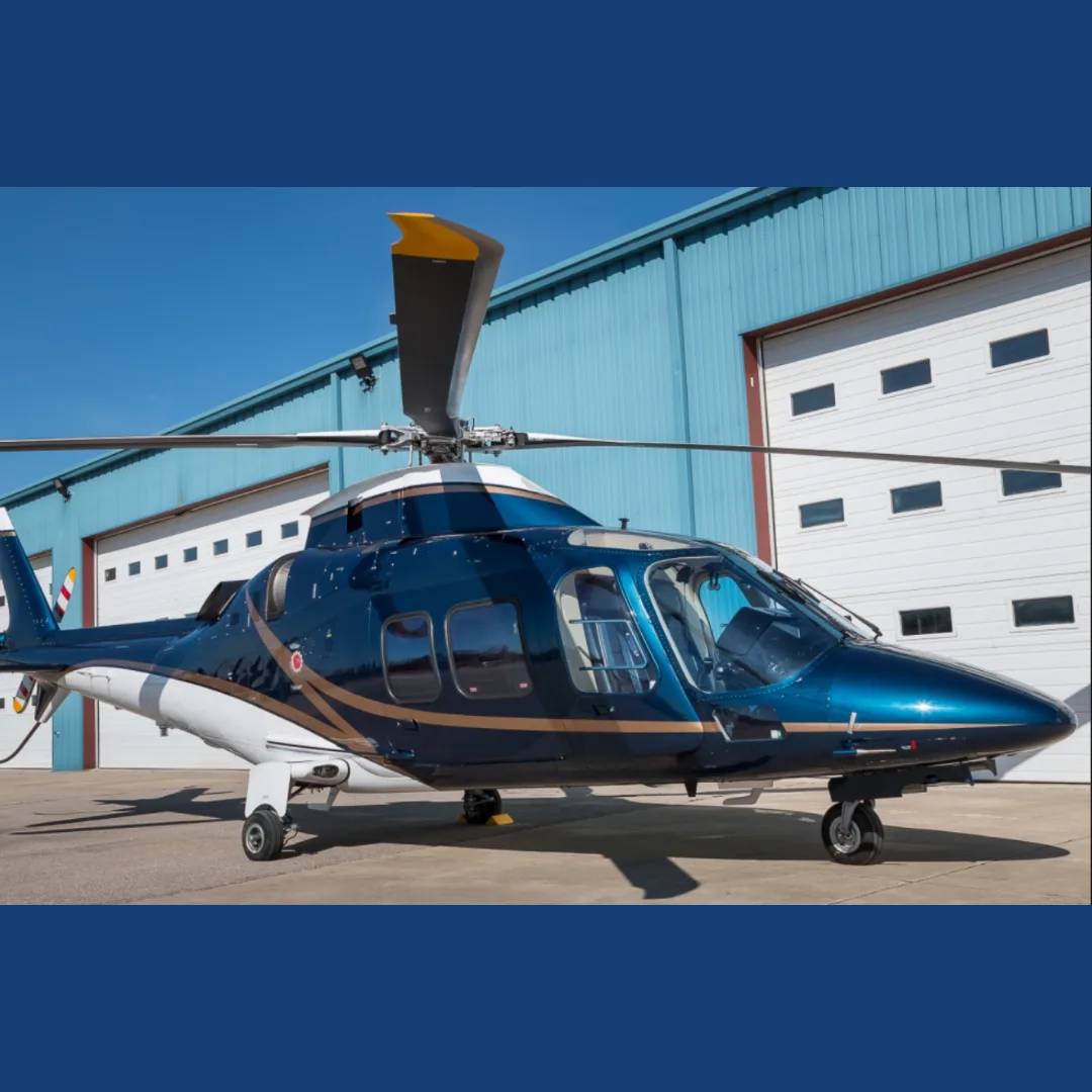 A dark blue and white luxury helicopter with gold accents is parked on an tarmac in front of a blue hangar.