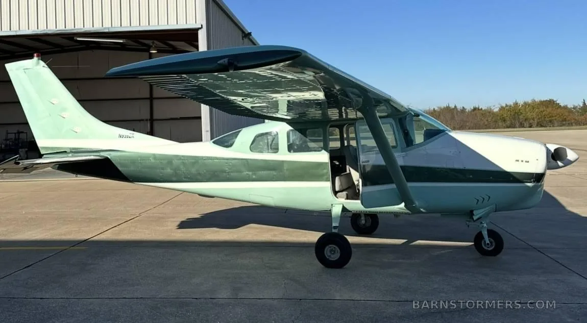 A light green and dark green Cessna 210 (N9982X) parked on an airfield next to a hangar.