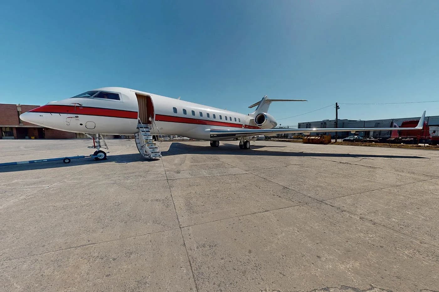A white private jet with a red stripe and open door stands on a concrete tarmac under a clear blue sky.