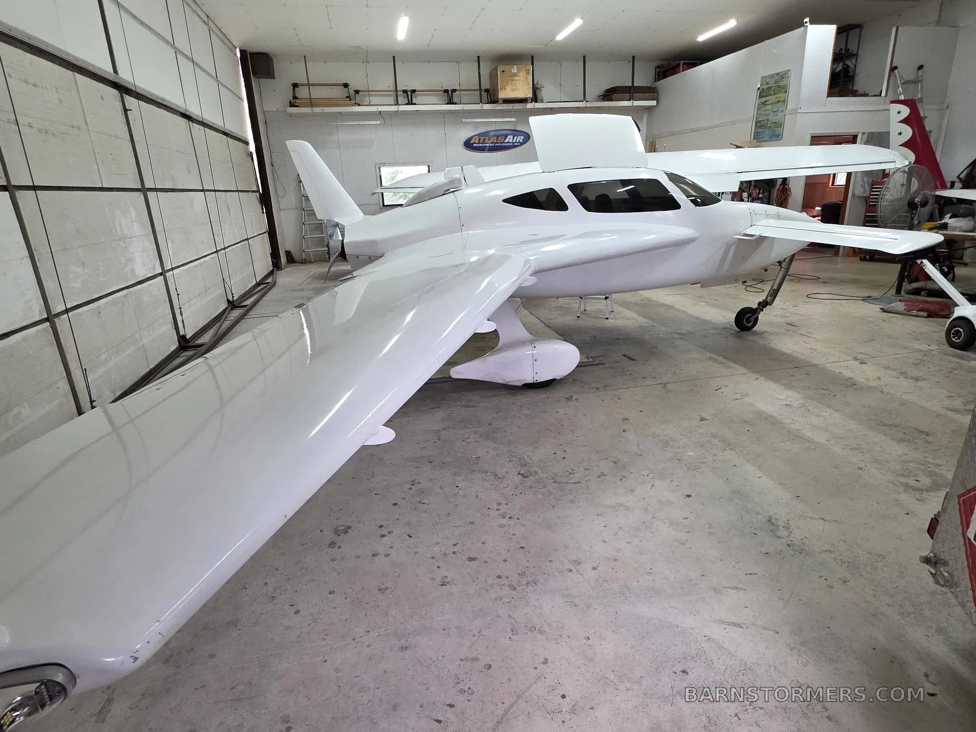 A white experimental aircraft with a pusher propeller is parked inside a hangar, viewed from the front-left.