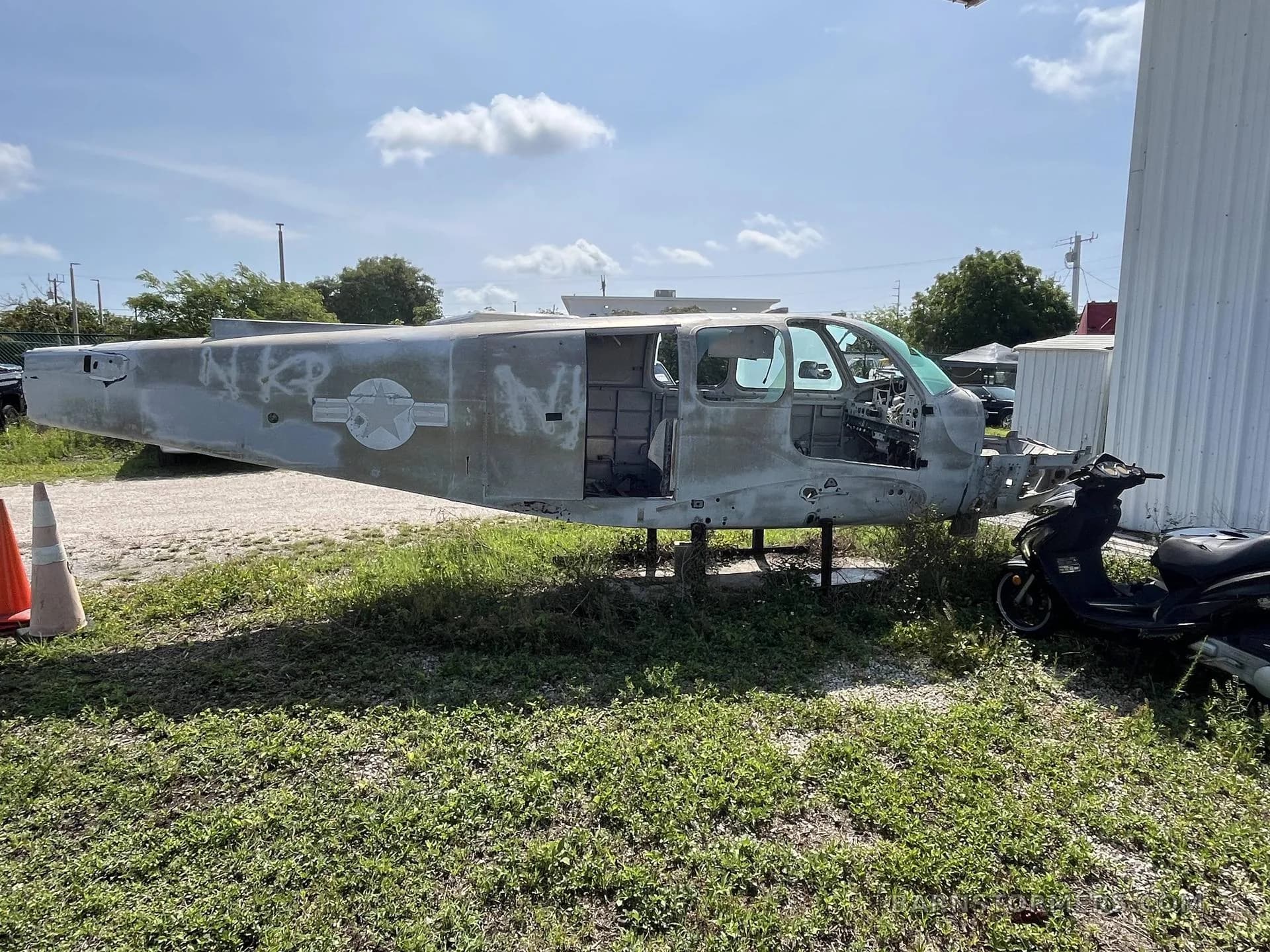 A stripped-down aircraft fuselage with a US military star insignia sits on grass next to a building under a sunny sky.