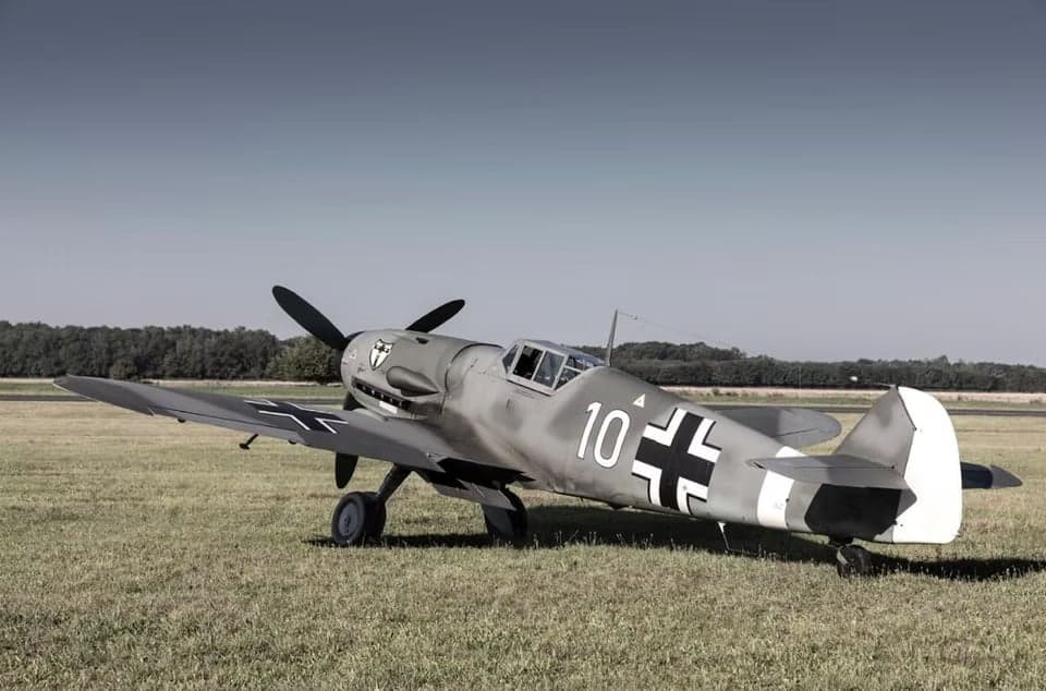 A gray Messerschmitt Bf 109 aircraft in flight over farmland.