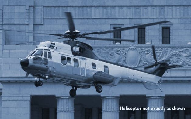 A blue-toned photo of a white and dark-colored helicopter hovering in front of a large stone building with ornate carvings.
