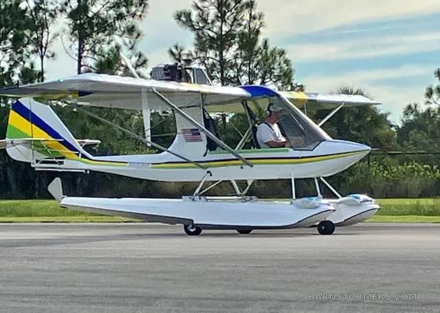 A white ultralight seaplane with green, yellow, and blue stripes, on floats on an asphalt surface.