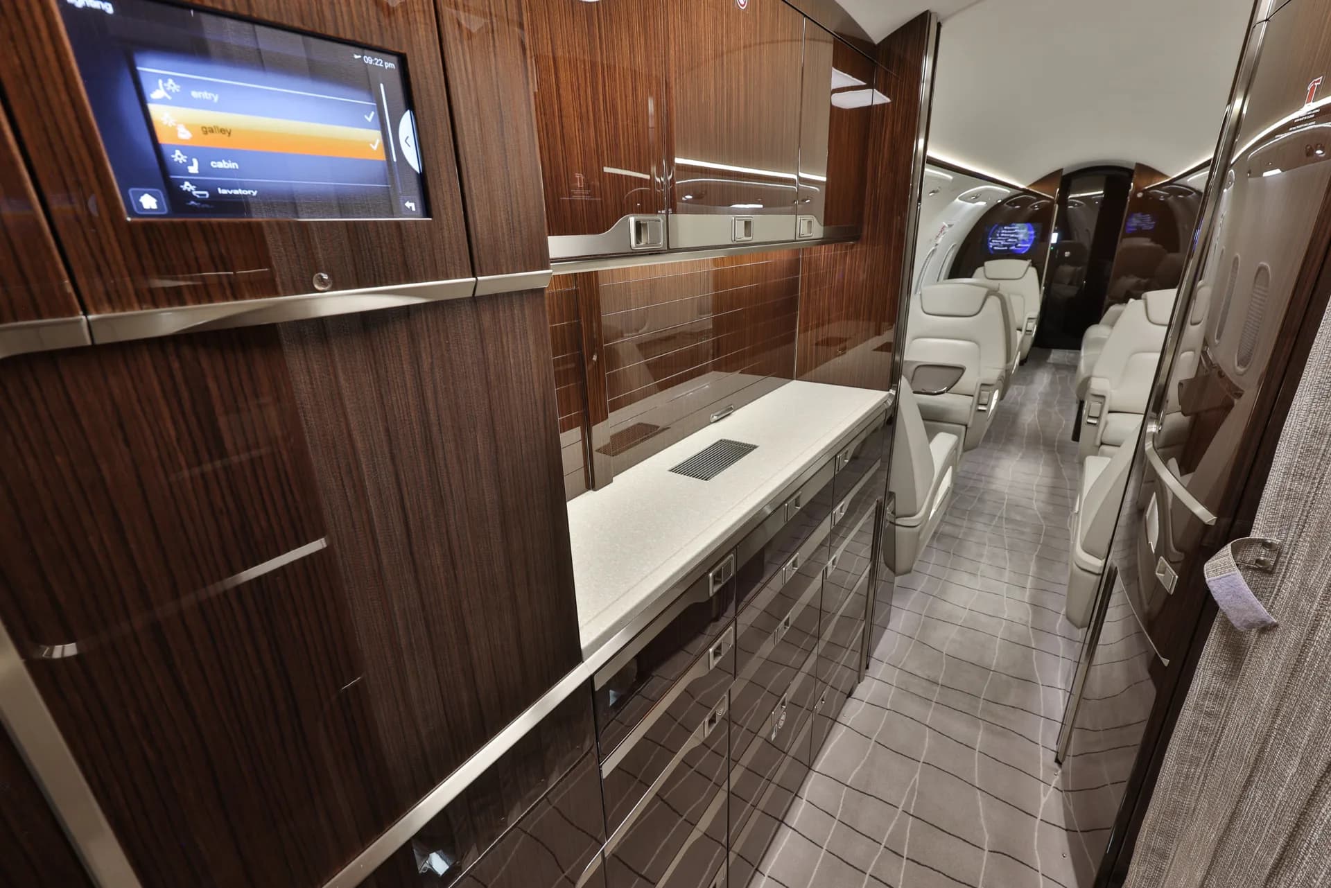 A modern aircraft galley with dark wood cabinetry, light countertops, and a control screen, leading into the cabin.