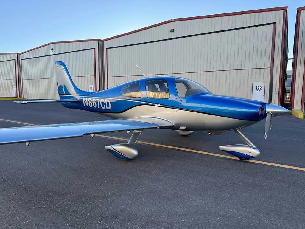 A blue and silver Cirrus SR22 aircraft with registration N867CD parked on an airfield in front of hangars.