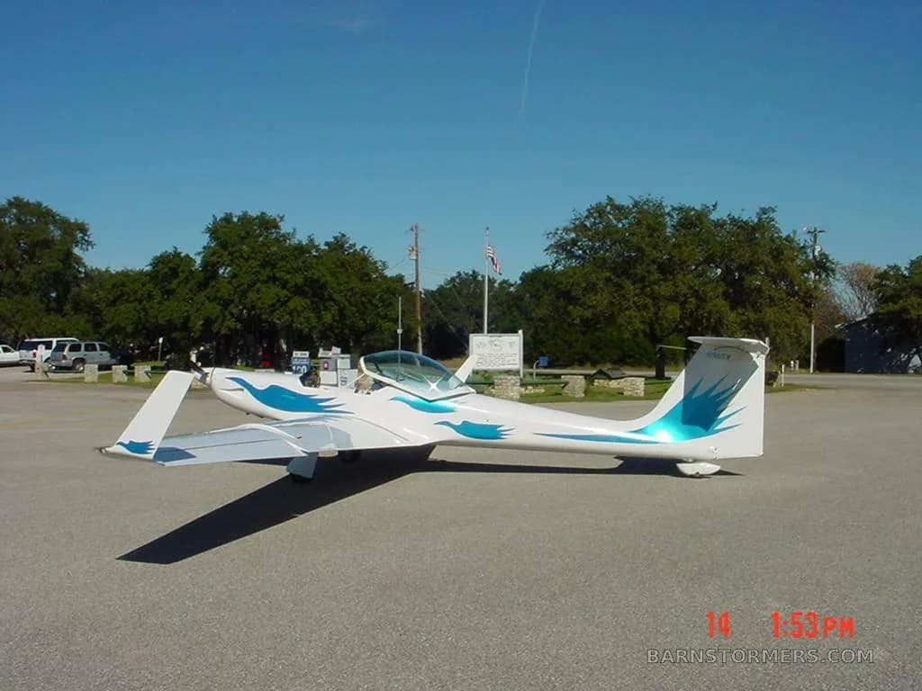 A white motorglider with blue wave decals on the fuselage and tail is parked on an asphalt lot under a clear blue sky.