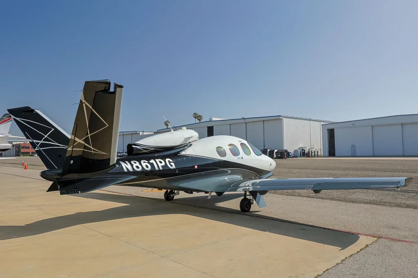 A Cirrus Vision Jet (N861PG) in white, black, and grey paint scheme parked on an tarmac with hangars in the background.