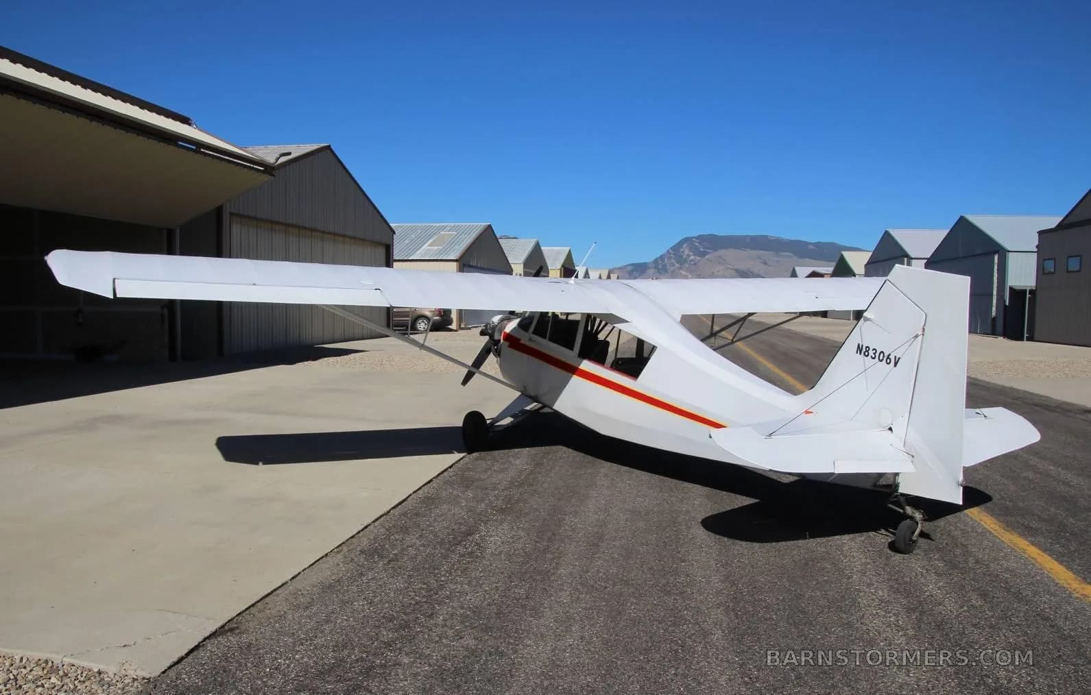 A white single-engine high-wing aircraft with red stripes and a red star on the cowl, seen from the rear-right, parked on an asphalt taxiway with mountains in the distance.