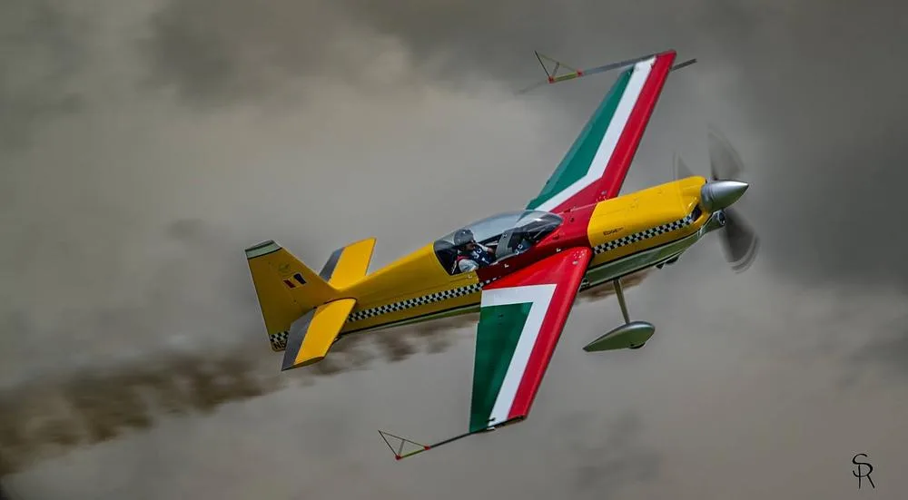 The N540TA aerobatic aircraft with red, green, yellow, and checkerboard livery flies against a cloudy sky.