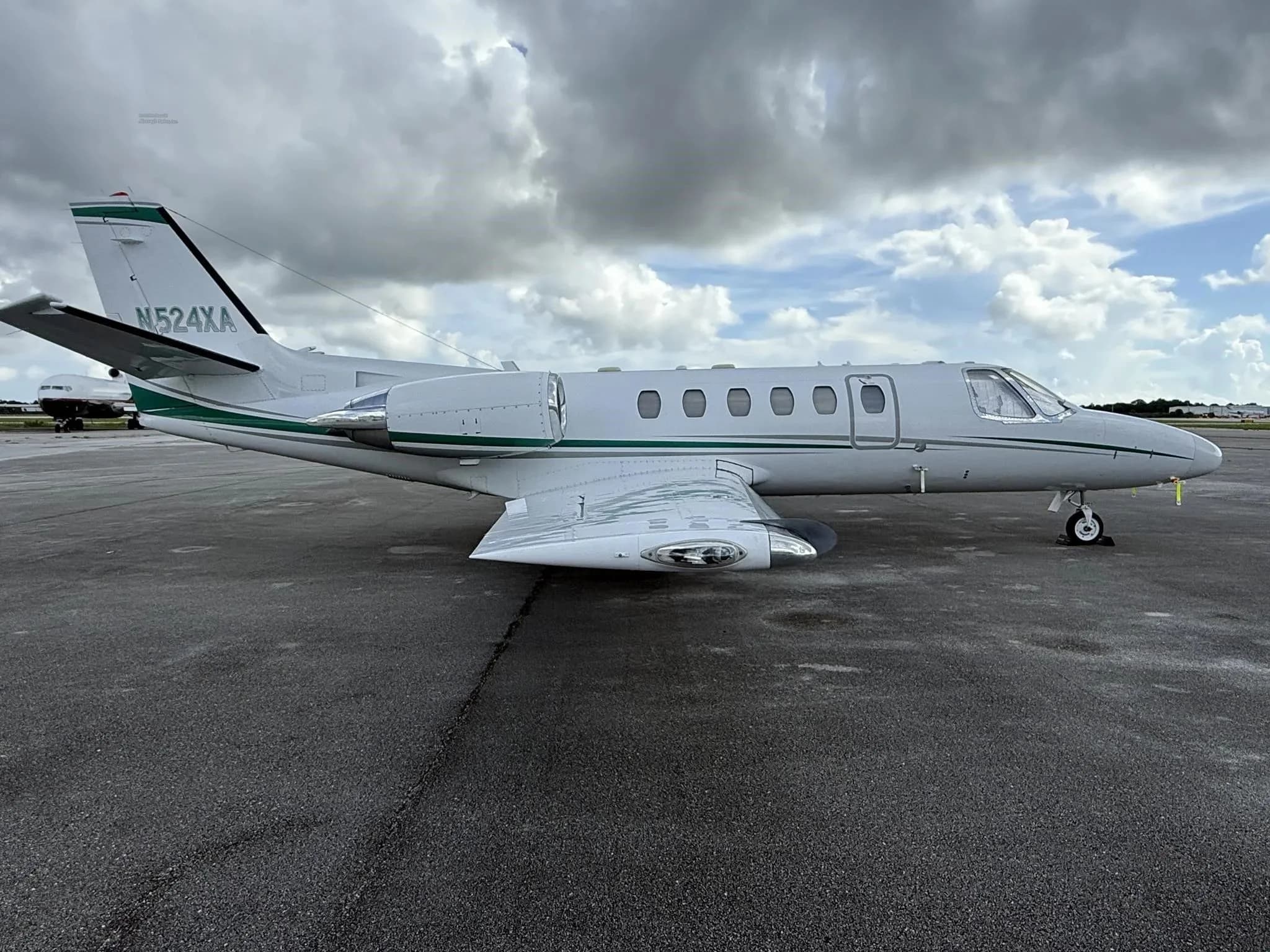 A side view of the white business jet N524XA with green stripes, parked on an airport tarmac under a cloudy sky.