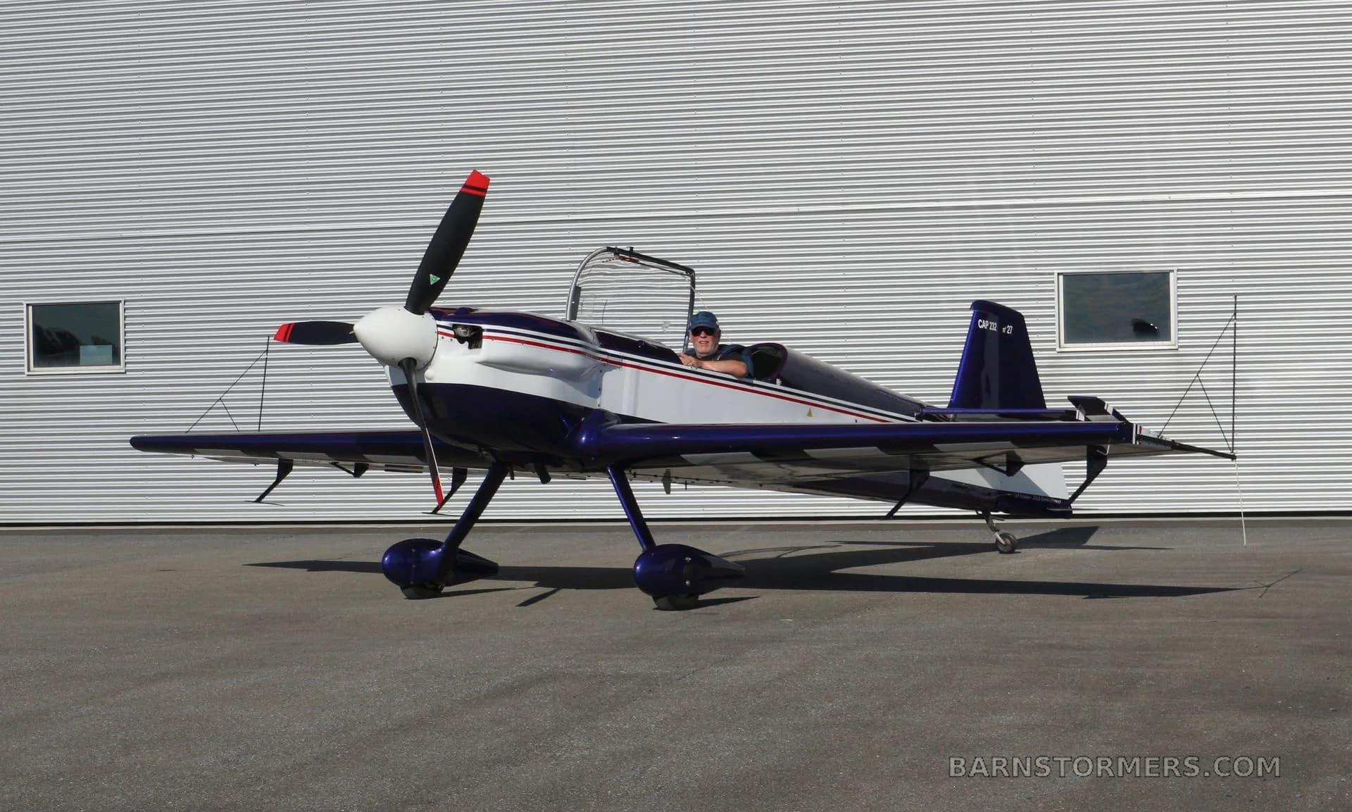 A blue and white CAP 10B aerobatic aircraft is parked on tarmac in front of a corrugated metal hangar, with a pilot in the cockpit.