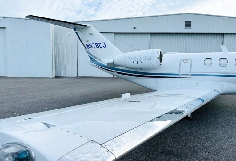 A rear view of a white Cessna Citation CJ3 (N578CJ) with blue and grey stripes, parked on an asphalt tarmac in front of a hangar.