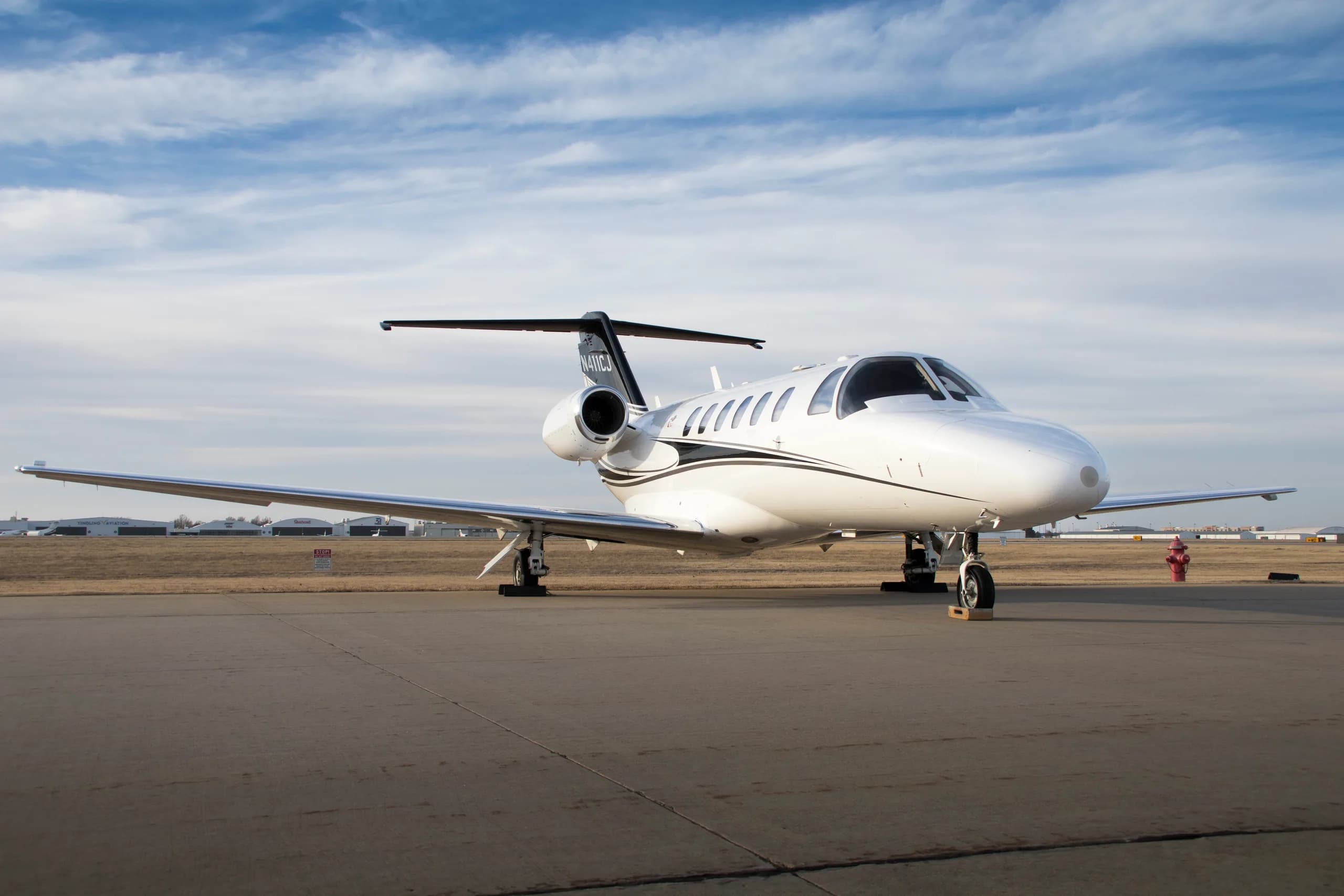A white Cessna Citation CJ3 private jet with black and gold stripes on the tail, registration N411CJ, parked on an airfield under a partly cloudy sky.