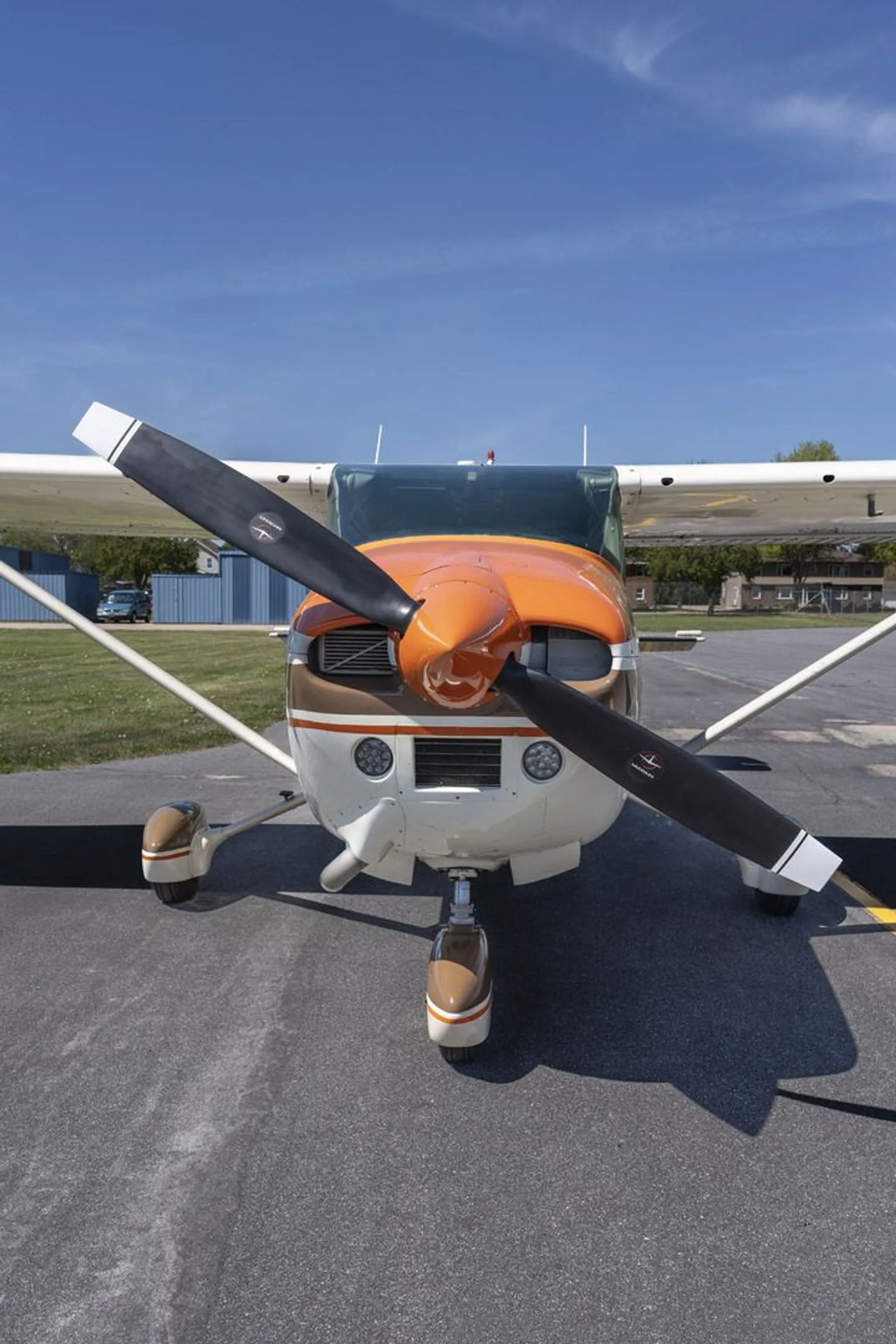 A head-on view of a white and brown Cessna 172 with an orange nose cone and propeller, parked on an asphalt ramp.